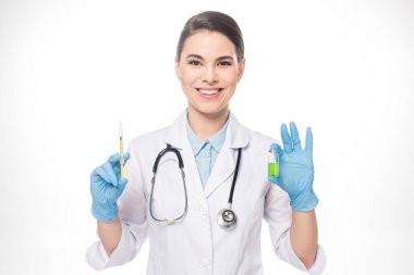 Attractive doctor smiling at camera while holding syringe and vaccine isolated on white clipart