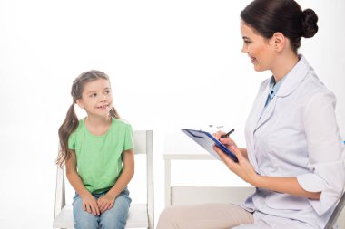 Smiling pediatrician with clipboard looking at kid on chair isolated on white clipart