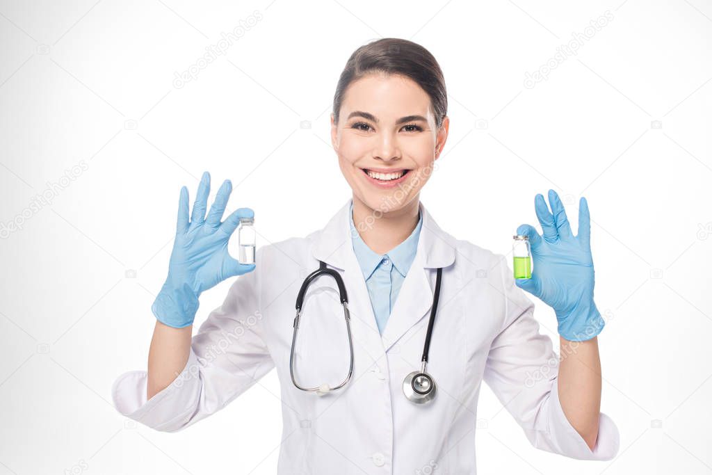 Beautiful doctor smiling at camera while holding jars with vaccines isolated on white