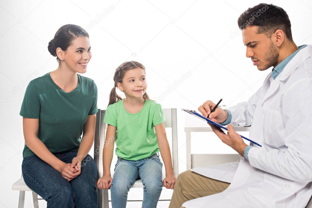 Smiling mother with daughter looking at pediatrician with clipboard isolated on white