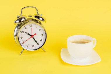 Sarı arka planda gümüş alarm saati ve bir fincan kahve.