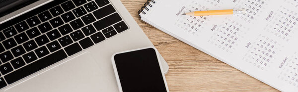 Panoramic shot of smartphone on laptop with calendar and pencil on wooden background