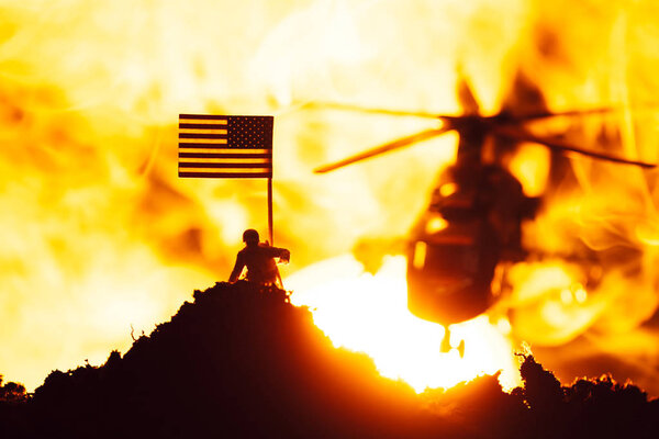 Battle scene with toy warrior near american flag and helicopter in fire with sunset at background