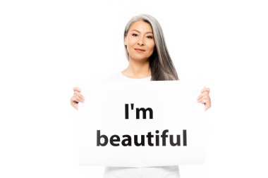 cheerful asian woman holding placard with i`m beautiful lettering isolated on white  clipart