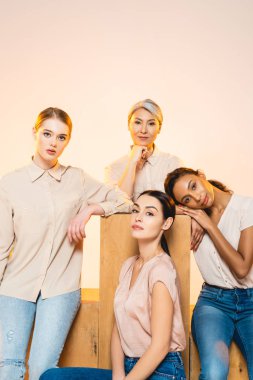four beautiful multicultural women with makeup looking at camera isolated on beige  clipart