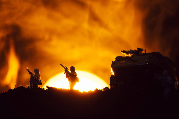Silhouettes of toy warriors and tank on battleground with sunset and fire at background