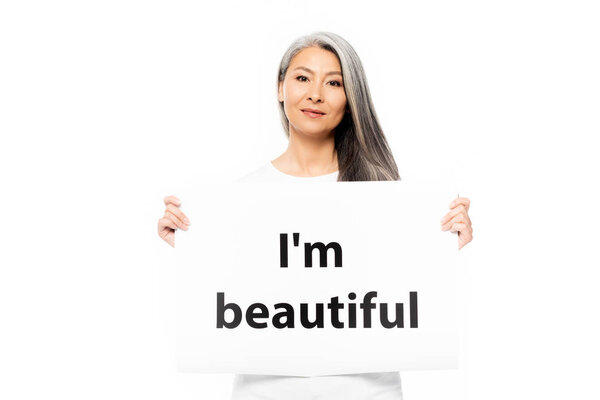 cheerful asian woman holding placard with i`m beautiful lettering isolated on white 