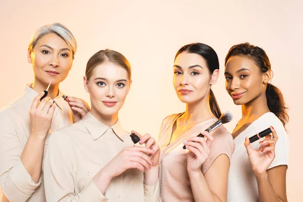 Heureuses Femmes Multiculturelles Tenant Des Cosmétiques Décoratifs Isolés Sur Beige — Photo