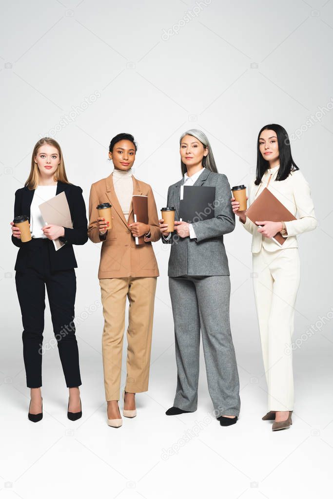 four multicultural businesswomen holding folders and paper cups while standing on white 