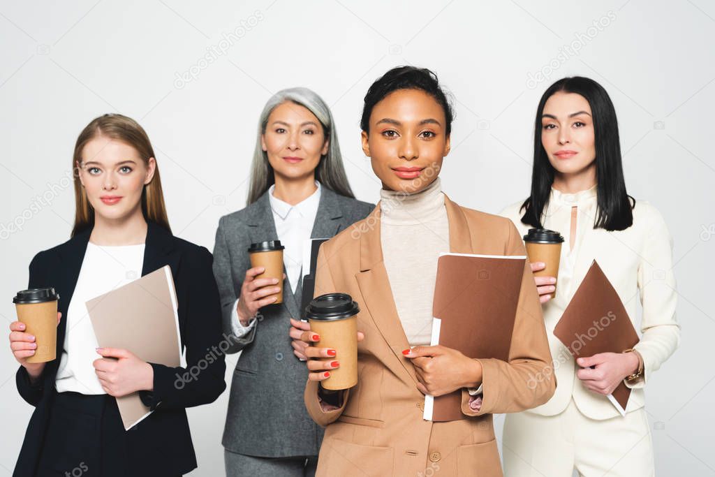 four attractive and multicultural businesswomen holding folders and paper cups isolated on white 