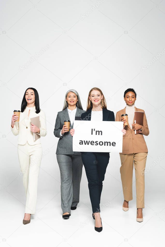 multicultural businesswomen holding folders and disposable cups near placard with i`m awesome lettering on white 