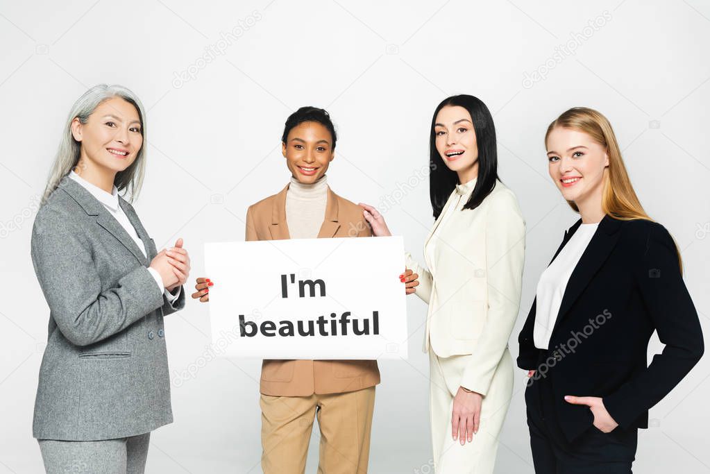 happy multicultural businesswomen standing near placard with i`m beautiful lettering isolated on white 