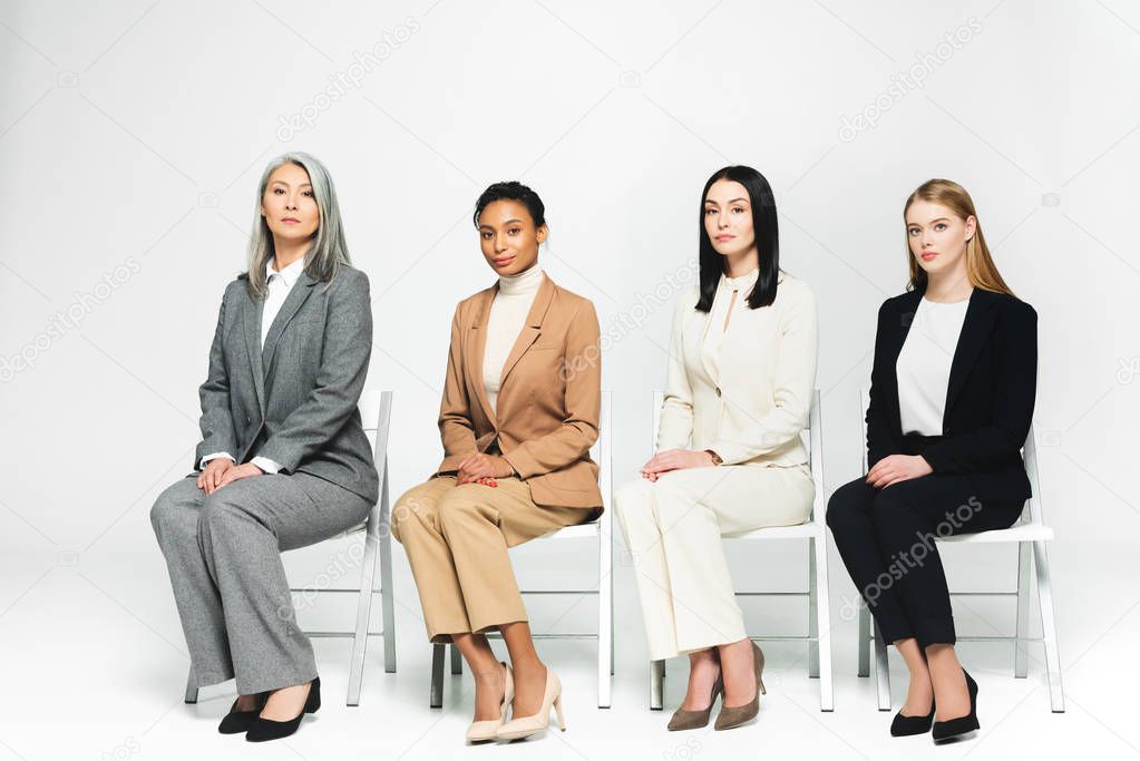 multicultural businesswomen in suits sitting on chairs and looking at camera on white 