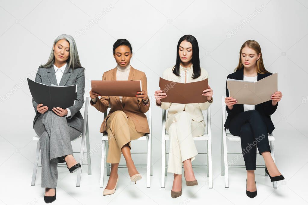 four multicultural businesswomen in suits sitting on chairs and looking at folders on white 