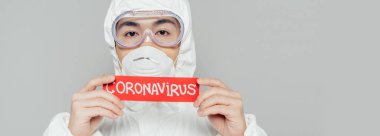 Tehlikeli madde giysisi ve solunum maskesi takmış Asyalı epidemiyolojistin koronavirüs yazıtlı uyarı kartı tutarken görüntüsü gri.