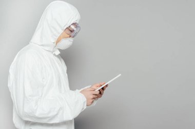 side view of asian epidemiologist in hazmat suit and respirator mask using digital tablet on grey background clipart