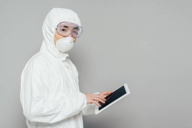 Tehlikeli madde giysisi ve solunum maskesi takmış Asyalı epidemiyoloji uzmanı elinde boş ekranlı dijital tablet tutarken gri arka plandaki kameraya bakıyor.