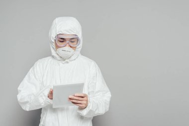 Tehlikeli madde giysisi ve solunum maskesi takmış Asyalı epidemiyoloji uzmanı gri arka planda dijital tablet kullanıyor.
