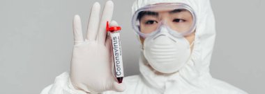 Tehlikeli madde giysisi ve solunum maskesi takmış Asyalı epidemiyolojistin panoramik görüntüsü gri üzerinde izole edilmiş kan örneğiyle test tüpünü gösteriyor.