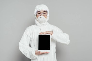 Tehlikeli madde giysisi ve solunum maskesi içinde Asyalı epidemiyoloji uzmanı gri arka planda boş ekranlı dijital tablet gösteriyor.