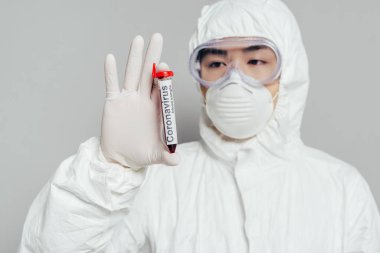 Tehlikeli madde giysisi ve solunum maskesi takmış Asyalı epidemiyoloji uzmanı gri arka planda kan örneği olan test tüpü gösteriyor.