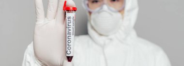 Tehlikeli madde giysisi ve solunum maskesi takmış Asyalı epidemiyolojistin seçici odak noktası gri panoramik iğne üzerinde kan örneğiyle test tüpü gösteriyor.