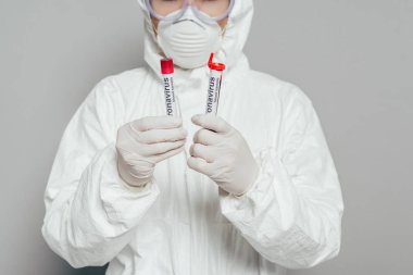Tehlikeli madde giysisi ve solunum maskesi takmış epidemiyoloji uzmanının gri arka planda kan örnekleri olan test tüpleri tutuşu.