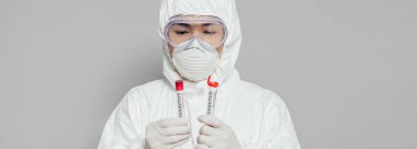 Asyalı epidemiyolojistin tehlikeli madde giysisi ve solunum maskesi içinde kan örnekleri içeren test tüpleri tutarken görüntüsü gri renkte.
