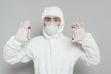 Tehlikeli madde giysisi ve solunum maskesi takmış Asyalı epidemiyoloji uzmanı gri arka planda kan örnekleri olan test tüplerini gösteriyor.