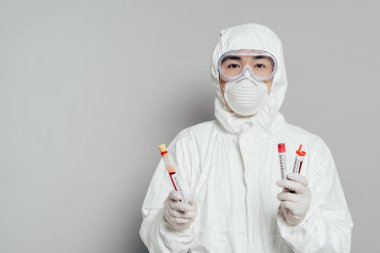 Tehlikeli madde giysisi ve solunum maskesi takmış Asyalı epidemiyoloji uzmanı gri arka planda kan örnekleri olan test tüpleri tutuyor.