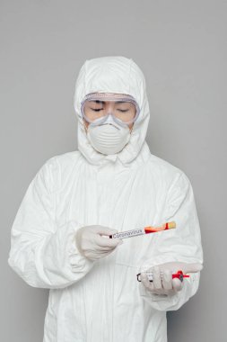 Tehlikeli madde giysisi ve solunum maskesi takmış Asyalı epidemiyoloji uzmanı gri arka planda kan örnekleri olan test tüpleri tutuyor.