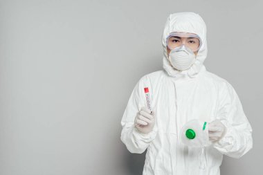 Tehlikeli madde giysisi içinde Asyalı epidemiyoloji uzmanı elinde solunum maskesi ve kan örneğiyle kameraya bakarken gri arka planda