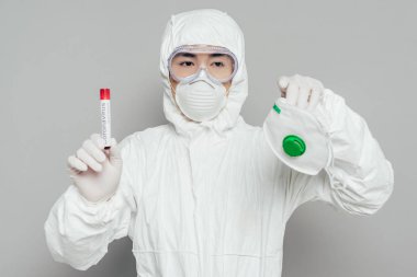 Tehlikeli madde giysisi içinde Asyalı epidemiyoloji uzmanı elinde solunum maskesi ve üzerinde kan örneği olan test tüpü tutuyor.
