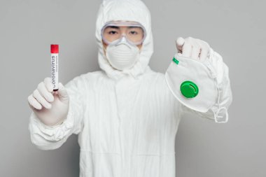 Tehlikeli madde giysisi içinde Asyalı epidemiyoloji uzmanı elinde solunum maskesi ve kan örneğiyle kameraya bakarken gri arka planda