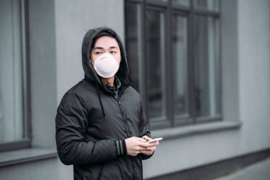 Solunum maskeli genç Asyalı adam akıllı telefon tutuyor ve sokakta dikilirken gözünü kaçırıyor.