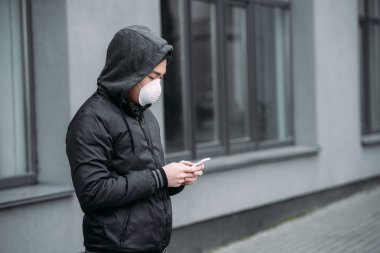 Solunum maskeli genç Asyalı adam sokakta dikilirken akıllı telefonla konuşuyor.