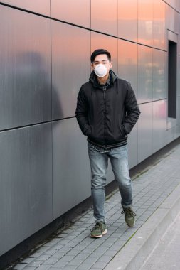young asian man in respirator mask looking at camera while walking along wall with hands in pockets