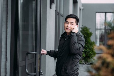 young asian man looking away while entering building and talking on smartphone
