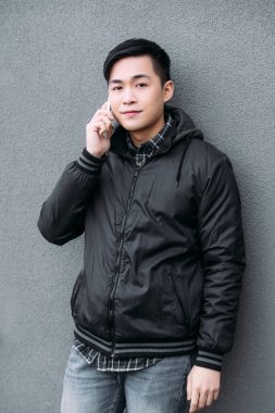 young asian man smiling while standing by grey wall, talking on smartphone and looking at camera
