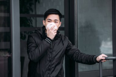 young asian man touching respirator mask while going out of building and looking away