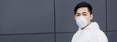 panoramic shot of asian epidemiologist in hazmat suit and respirator mask looking at camera while standing near wall