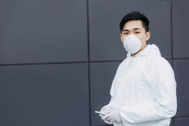 asian epidemiologist in hazmat suit and respirator mask holding smartphone and looking at camera while standing near wall