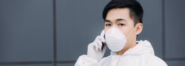 panoramic shot of asian epidemiologist in hazmat suit and respirator mask talking on smartphone and looking at camera