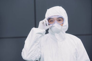 asian epidemiologist in hazmat suit and respirator mask looking at camera while standing outside and touching goggles