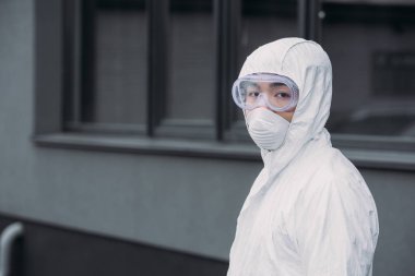 asian epidemiologist in hazmat suit and respirator mask looking at camera while standing on street