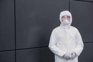asian epidemiologist in hazmat suit and respirator mask looking at camera while standing near wall