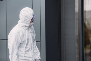 epidemiologist in hazmat suit standing outside and looking in window of building