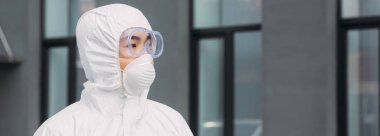 panoramic shot of asian epidemiologist in hazmat suit and respirator mask looking away while standing on street near building
