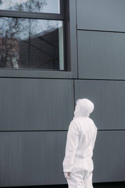 back view of epidemiologist in hazmat suit standing outside and looking in window of building
