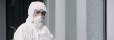 panoramic shot of asian epidemiologist in hazmat suit and respirator mask looking at camera while standing outside